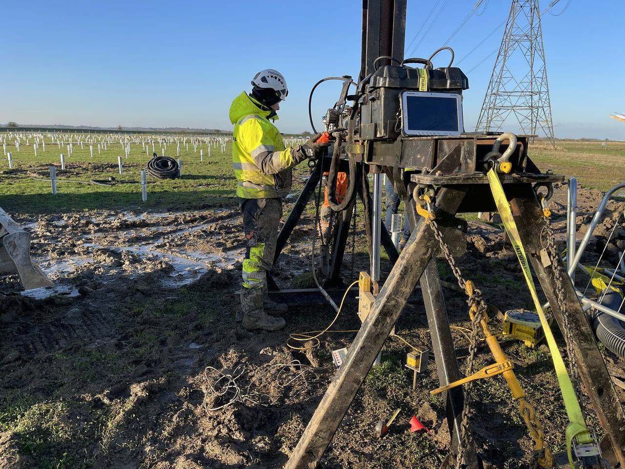 Helicasol crew member operating a test-piling rig on site, installed piles and transmission lines in the background